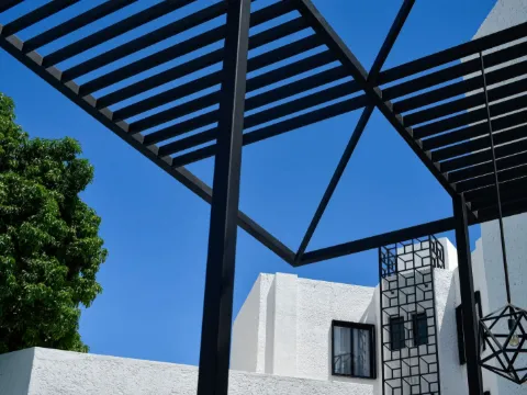Pergola à poutres noires fines et lampe géométrique devant une maison blanche sous un ciel bleu
